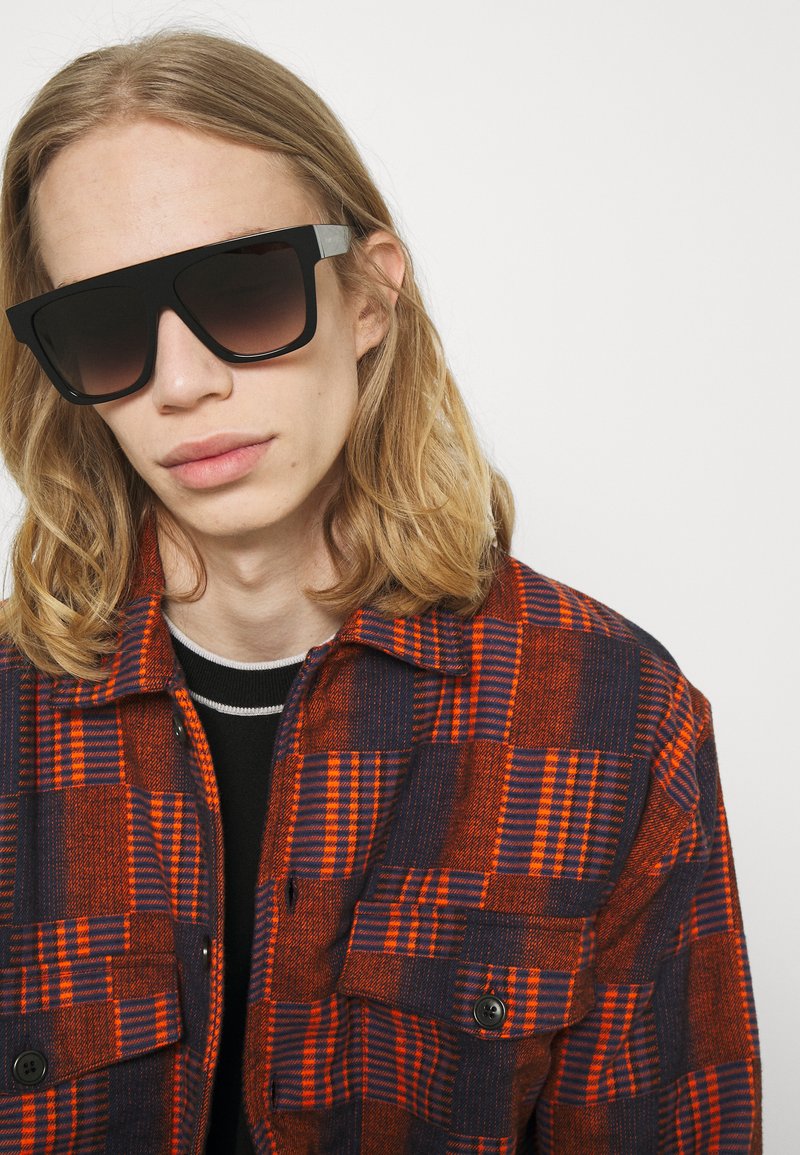 Schwarze übergroße Sonnenbrille mit getönten Gläsern. Rote und marineblaue Karojacke mit strukturiertem Stoff und knöpfbaren Fronttaschen.