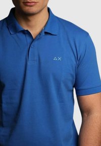 Polo blu realizzato in tessuto liscio, con colletto classico, maniche corte e un logo sottile in una tonalità più chiara sul petto.