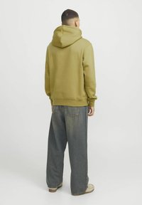 Olivgrüne Baumwollkapuzenjacke mit großer Kapuze, gerippten Bündchen und Saum; kombiniert mit locker sitzenden, ausgewaschenen blauen Jeans und beigen Schuhen.