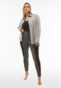 Camicia grigia chiaro button-up indossata sopra una maglietta nera, abbinata a leggings neri e aderenti con motivo. I leggings presentano un design a scacchi e pannelli laterali.