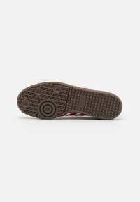 Brown rubber outsole with a textured gripping pattern, featuring circular accents and a logo. The shoe has red stripes along the side.