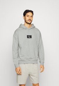 Felpa grigia con cappuccio e patch logo sul davanti, realizzata in tessuto morbido. Presenta una vestibilità comoda, polsini e orlo a coste.