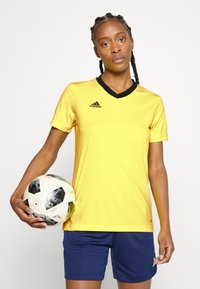 Geltona sportinė maikutė su juodais V formos akcentais, derinama su mėlynomis šortais. Modelis laiko futbolo kamuolį, papuoštą juodai baltu geometriniu raštu.