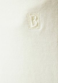Krémová, texturovaná tkanina se malým vyšitým čtvercovým logem s písmenem "B". Design je jednoduchý a minimalistický.