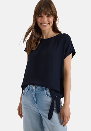 Mujer sonriente con el pelo castaño liso, vistiendo una camiseta holgada azul marino de manga corta con lazo lateral y vaqueros azul claro sobre un fondo liso.