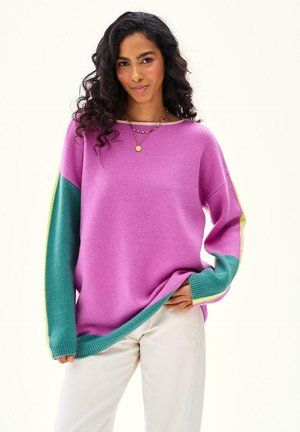 Kobieta o kręconych włosach ubrana w luźny fioletowy sweter z turkusowymi i żółtymi rękawami, beżowe spodnie oraz kolorowe naszyjniki z koralików.
