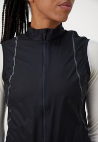 Schwarze ärmellose Jacke mit hohem Kragen, Frontreißverschluss, texturiertem Stoff und kontrastierenden hellgrauen Nähten.