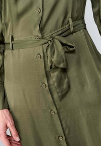 Robe vert olive en tissu lisse, avec un devant à boutons, une ceinture nouée à la taille et des manches longues.