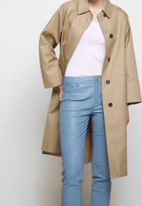 Cappotto impermeabile beige con colletto e bottoni neri, abbinato a pantaloni in pelle blu chiaro con un dettaglio della tasca frontale.