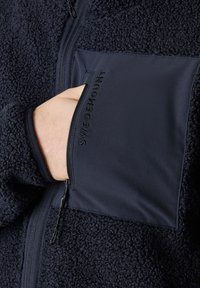 Giacca in pile blu navy con un corpo testurizzato e morbido. Presenta una tasca sul petto in tessuto liscio con la scritta "SWEDEMOUNT" impresso. Chiusura frontale con zip.