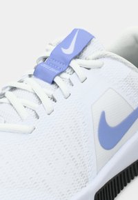 Hvit treningssko med teksturert overdel, lilla detaljer og en fremtredende Nike-logo. Har tradisjonell snøring og polstret krage.