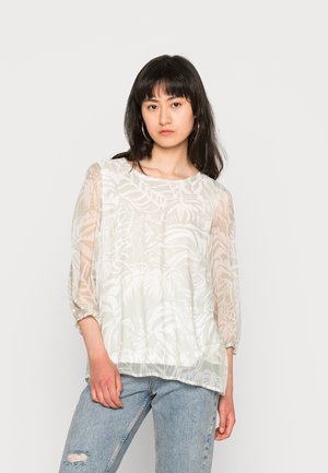 Blusa branca, transparente, com um padrão de folhas, decote redondo e mangas 3/4, combinada com jeans claros, desgastados. Textura suave.