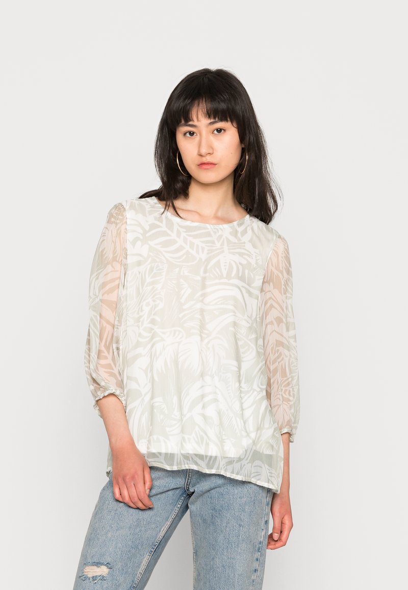 Blusa branca, transparente, com um padrão de folhas, decote redondo e mangas 3/4, combinada com jeans claros, desgastados. Textura suave.
