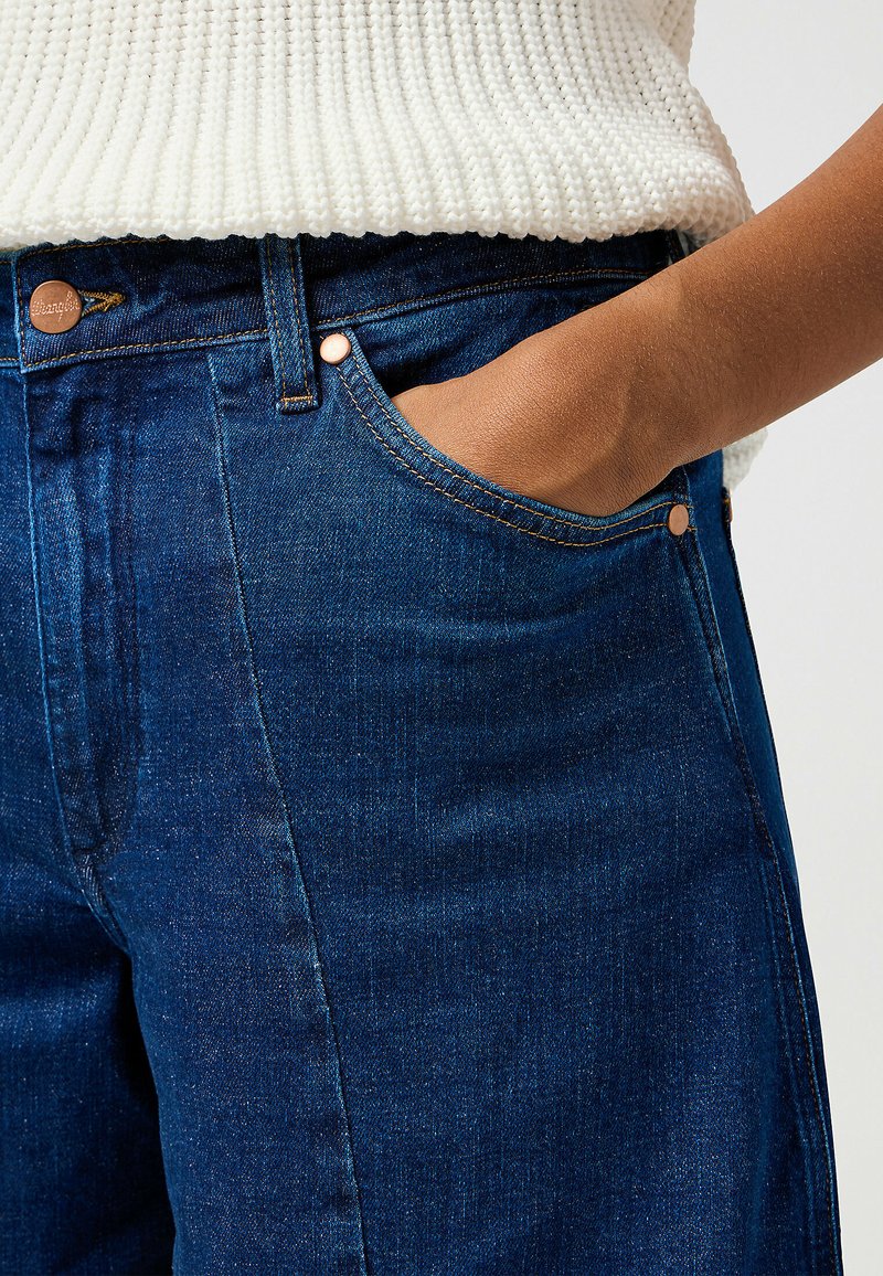 Jean en denim bleu foncé avec taille haute, poche avant et accessoires en ton cuivre, avec des coutures visibles sur les coutures.