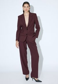 Blazer ajusté bourgogne avec revers crantés, fermeture à un bouton et pantalon large assorti ; associé à des escarpins noirs à bout pointu.
