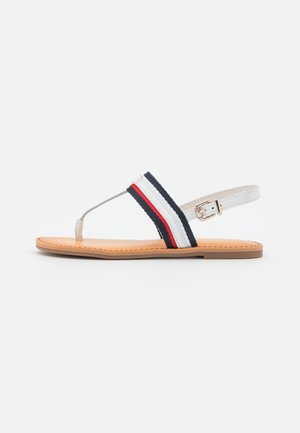 Flad sandal med tanfarvet sål, hvid tårem, justerbar hvid ankelrem og navyblåt, hvidt og rødt stribet stofbånd over foden.