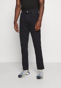 Jeans de mezclilla negra con corte recto, diseño de cinco bolsillos y cierre de botón. Combinados con zapatillas deportivas grises. Construcción sencilla.