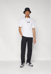 Camiseta blanca con estampado gráfico, pantalones cargo negros con bolsillos, sombrero tipo bucket negro y zapatillas en blanco y negro. Pose de pie sobre un fondo liso.