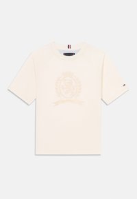 Krämfärgad t-shirt med en präglad sköld med en lejon, omgiven av lagerblad och ett banderoll, presenterad på en enfärgad bakgrund.
