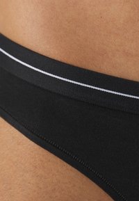 Fekete bikini alsó sima textúrával, vékony fehér díszcsíkkal a derékvonal mentén. A dizájn minimalista és testhez simuló.