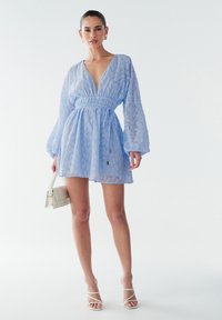 Svetlo modra obleka z dolgimi voluminoznimi rokavi, teksturirano tkanino, globokim V-izrezom, stisnjenim pasom in polprosojnim zaključkom. Dodatki vključujejo majhno torbico.