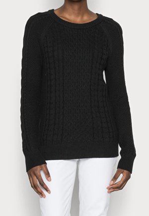 Person wearing a black cable-knit sweater with long sleeves and white pants, standing against a plain light background.