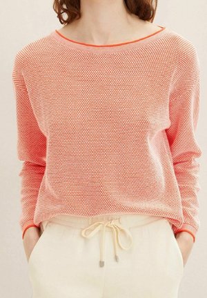 Personne portant un pull ample rose et blanc texturé avec une bordure orange et un pantalon crème à cordon avec les mains dans les poches.