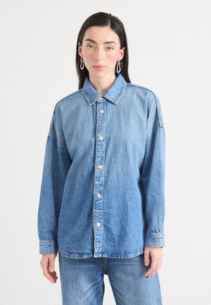 Een jonge vrouw staat tegen een lichte achtergrond, gekleed in een blauwe spijkerblouse met lange mouwen, dichtgeknoopt, en eenvoudige oorbellen.