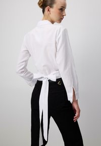 Camicia bianca a maniche lunghe con nodo dietro, dotata di colletto, fessure laterali e polsini con bottoni, abbinata a pantaloni neri. Tessuto dalla texture liscia.