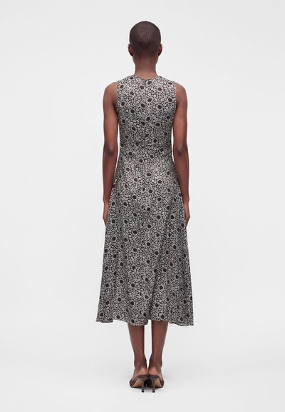 Femme aux cheveux courts portant une robe midi à fleurs noire et blanche sans manches et des sandales à talons noires, debout de dos.