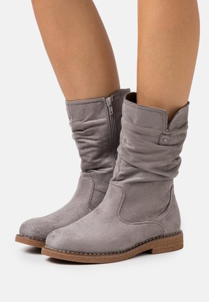 Femme portant des bottes en daim gris mi-mollet avec tige souple, fermeture éclair latérale et semelle en caoutchouc marron sur un fond clair uni.