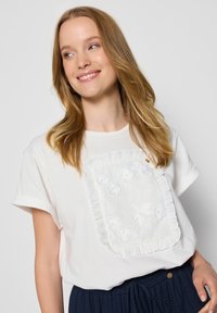 Biała koszulka z bawełny z falbaniastą, haftowaną kieszonką z motywem kwiatowym. Krótkie rękawy i okrągły dekolt, zestawiona z ciemnymi spodniami.