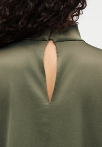 Blouse vert olive avec un col montant, présentant une petite ouverture en trou de serrure dans le dos et une texture lisse et mate.