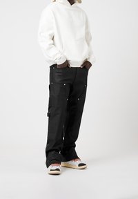 Sweat à capuche blanc avec une poche avant, pantalon cargo noir avec plusieurs poches et boutons-pression, et baskets avec des accents colorés.