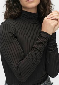 Musta ribattua pitkähihaista poolopaitaa, jossa on viivamaista tekstuuria. Koon mukaan istuva malli, jossa on rypytetyt hihansuut, ja se on valmistettu sileästä, joustavasta kankaasta.