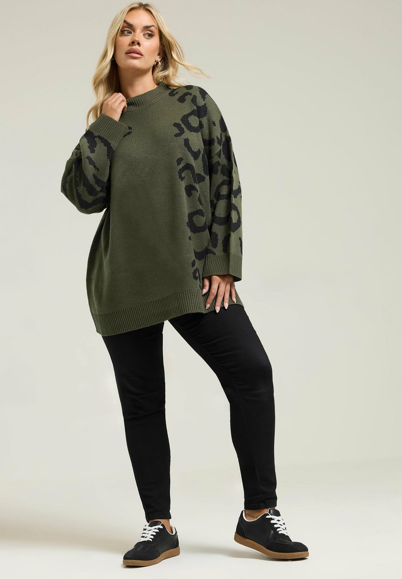 Khaki zielony, oversize'owy sweter z dzianiny w czarną panterkę, z prążkowanymi mankietami i dołem, połączony z czarnymi dopasowanymi spodniami i czarnymi trampkami.