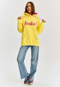 Žltá mikina s kapucňou a ružovým grafickým nápisom "¡hola!", predné vrecko a nastaviteľná šnúrka na kapucni, spojená so svetlo modrými džínami a viacfarebnými teniskami.