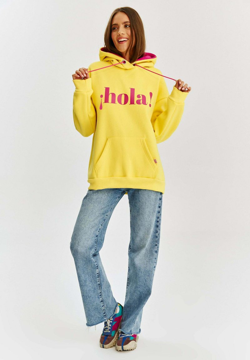 Žltá mikina s kapucňou a ružovým grafickým nápisom "¡hola!", predné vrecko a nastaviteľná šnúrka na kapucni, spojená so svetlo modrými džínami a viacfarebnými teniskami.