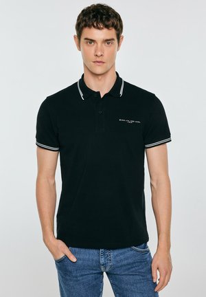 Młody mężczyzna z krótkimi kręconymi włosami, ubrany w czarną koszulkę polo z białym wykończeniem i niebieskie dżinsy, stojący z jedną ręką w kieszeni.