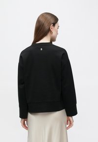 Μαύρο φούτερ με χαλαρή εφαρμογή, στρογγυλό λαιμό, ραμμένους ώμους και μικρό λογότυπο στο πίσω μέρος. Με λεία ύφασμα και ριμπ μανσέτες.