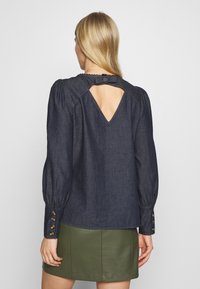 Blusa in denim blu scuro con aperture a occhio di bue sulla schiena, maniche a sbuffo e dettaglio con bottoni dorati ai polsini. Tessuto morbido e vestibilità rilassata.