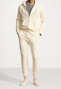 Mężczyzna w jasnobeżowej bluzie z zamkiem, pasujących joggerach, beżowych skarpetach i jasnobeżowych sneakersach, stojący z rękami w kieszeniach.