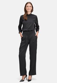 Schwarze Satinbluse mit hohem Kragen und Dreiviertelärmeln, kombiniert mit hochtaillierten schwarzen Hosen mit Falten und Seitentaschen.