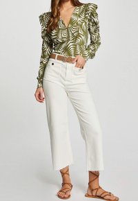 Blouse verte à manches longues avec motif de feuilles blanches, associée à un jean blanc court. Ceinture beige avec détails métalliques et sandales marron à lanières.
