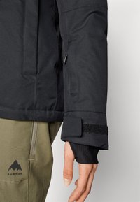 Must musta isolatsiooniga jope varrukas, millel on Velcro rihm, koos oliivroheliste pükstega, millel on mägi logo. Sile tekstuur, funktsionaalne disain.