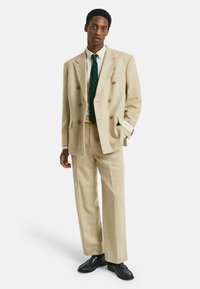 Homme portant un costume beige à double boutonnage avec une cravate rayée verte, une chemise blanche et des chaussures habillées noires, se tenant avec une main dans la poche.