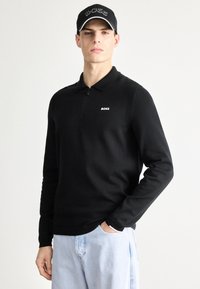 Črna polo majica z dolgimi rokavi, polovičnim zadrgo in ovratnikom, z majhnim belim logotipom. Opravljena s svetlo modrimi kavbojkami.