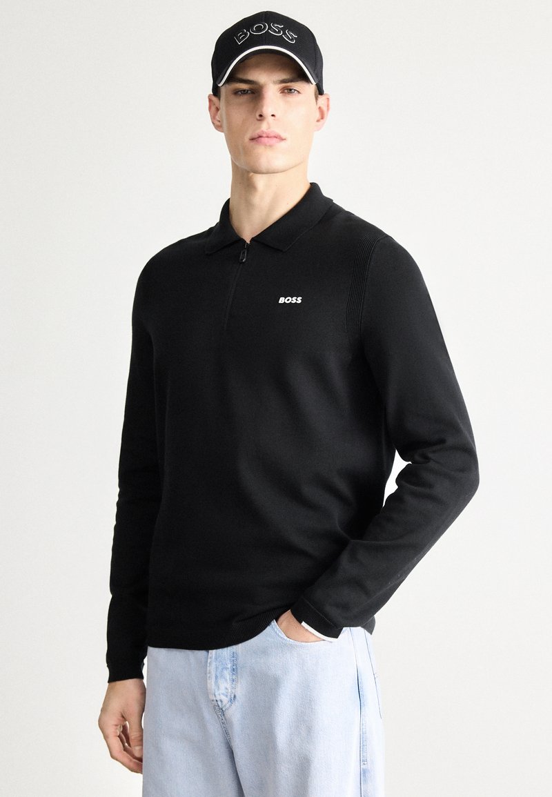 Črna polo majica z dolgimi rokavi, polovičnim zadrgo in ovratnikom, z majhnim belim logotipom. Opravljena s svetlo modrimi kavbojkami.