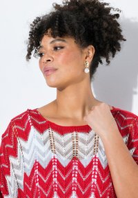Top a righe rosse e bianche in maglia con scollatura ampia e lavorazione a rete. Presenta un modello che indossa orecchini a perla e ha un'acconciatura riccia.