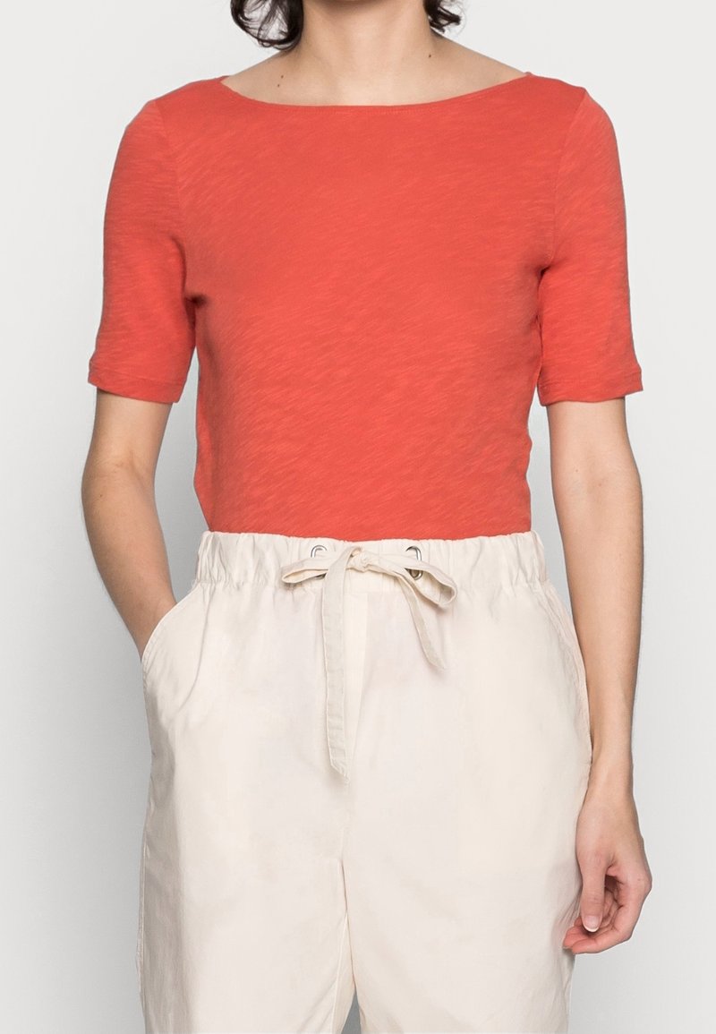 Top rosso a maniche corte con scollatura a barca, realizzato in tessuto texturizzato. Abbinato a pantaloni beige morbidi dotati di cordoncino in vita.